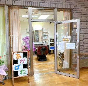 介護施設の玄関エリア。ガラス扉とレンガ壁、案内表示が見られます。