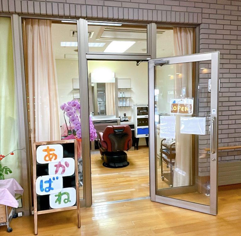 介護施設の玄関。ガラス戸に「あかばね」の看板、紫色の花が飾られている。